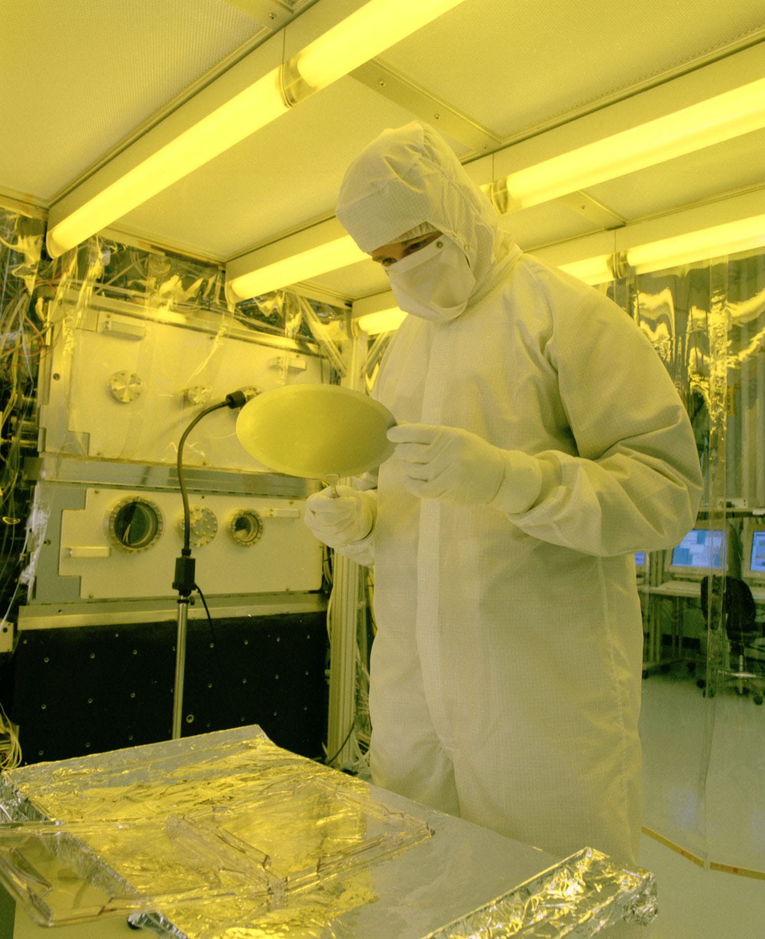 An engineer in a cleanroom suit inspects a silicon wafer under yellow safe lights